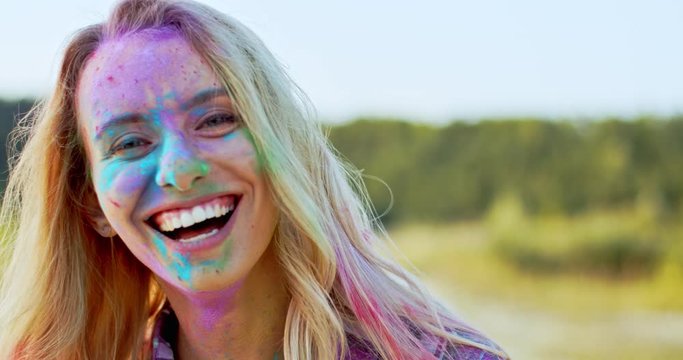 Portrait of the blond young and beautiful Caucasian woman looking straight to the camera and smiling cheerfully while being in colorful spots of paints outside. At the holi festival.