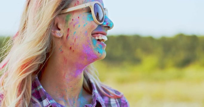 Close up of the young blond beautiful Caucasian woman in sunglasses smiling and looking at the side while her face being in colorful paints as she celebrating Holi, Indian holiday.