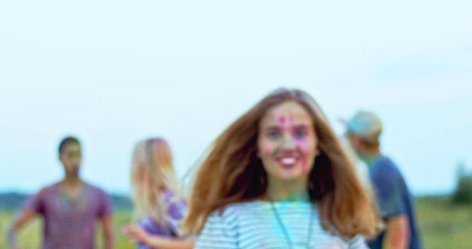 Portrait of the attractive young Caucasian joyful girl coming closer to the camera and smiling to it while posing with many friends celebrating holi fest on the background. Outdoors.