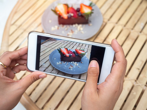 Woman's hand taking a photo of delicious chocolate tart on ceramic dish by smartphone in cafe.