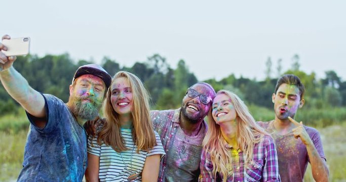 Group of young cheerful multiethnic male and female friends in colorful paints powder smiling to the smartphone camera while taking selfie photo outsideat the holi fest.