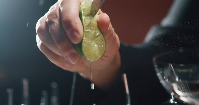 Close up of a professional bartender is squeezing a lime while preparing an alcoholic cocktail with ice cubes to customers at the bar or disco club.