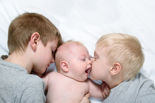 Two older brothers tenderly kiss and hug the younger child on a white bed. Happy family