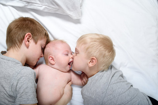 Two older brothers tenderly kiss and hug the younger child on a white bed. Happy family