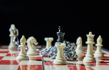 Black king is imprisoned on the chess board with chains and is surrounded by white pieces