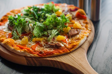 Fresh pizza with meat, pineapples and salad served on a board on black wooden table