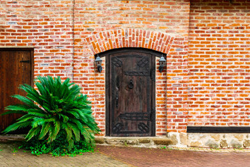 A closed old wooden massive door with a knock on a doorknob in the form of a twisted ring, vintage lanterns hang on both sides of a red brick wall and a green shrub. Background, copy space.