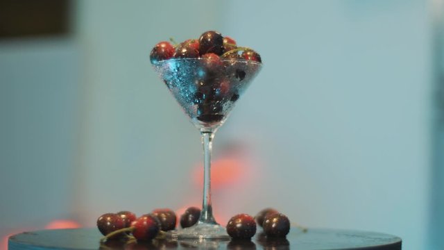 Camera is dynamically rotating around one coctail glass full of dark-red berries placed on round-shaped table in room with dark-blue walls and illumination lights.