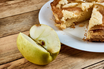 Naklejka na ściany i meble Sliced piece of green apple next to a baked apple pie on a white plate on a wooden background