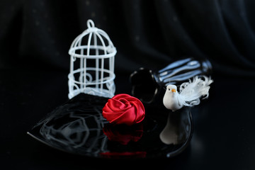 Solemn festive table setting in dark key with use black dishes, decorations of red roses and white birdcage. Symbol of love and passion.