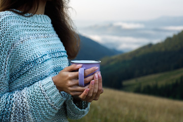 Closeup photo of cup with coffee in traveler's hand. Young woman drinks a hot drink from a cup and enjoys the scenery in the mountains