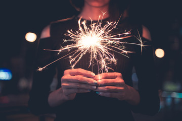 Lady standing in center holding sparklers stick on fire with both hands, have light bokeh of light bulb and car moving at night time - toning