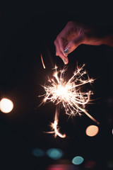 Woman's hand holding sparklers stick on fire from top drop down, big flame spread long light with bokeh of light bulb at night time on black background