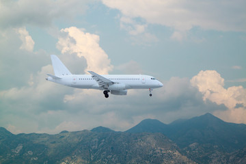white passenger plane landing against mountains with white clouds on blue sky background.  Landscape with airplane, mountains, sea and blue sky with white clouds in summer. Business travel in Europe.