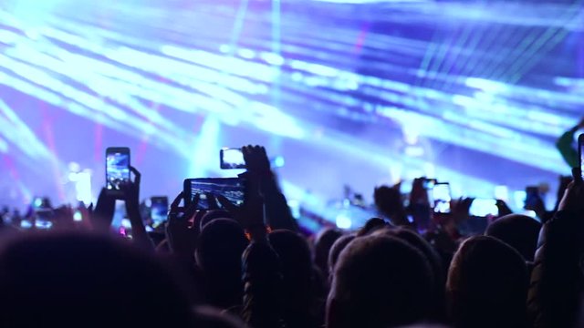 Phones in the hands of a crowd of viewers take videos and photos at a music festival. Spectators filmed the artist on the phone during a performance on stage. Laser show on live music concert.