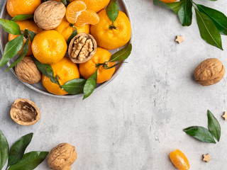 Fresh bright tangerines (oranges) with green leaves and walnuts on gray plate on pullover background, cozy winter mood, top view, frame, template layout