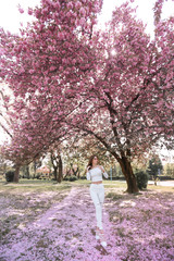 Wide angle photo of a woman in white jeans in a pink garden with trees dotted with cherry blossoms.