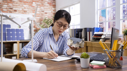 Fototapeta na wymiar pretty female architect working on notebook. asian korean woman engineer in workplace planning architectural project with green energy. elegant girl worker writing new ideas note with 3D house model