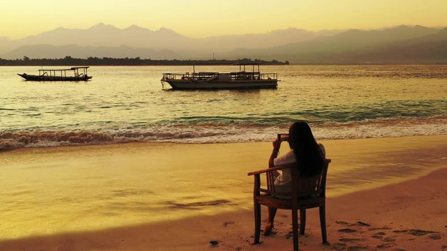 young beautiful woman sitting in the evening at sunset on the sady beach by the sea and taking a landscape photo on her smartphone.