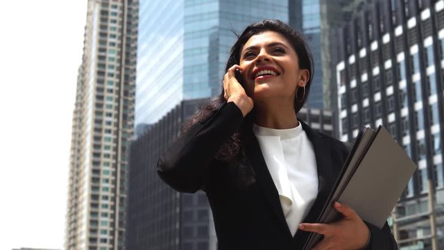 Happy young indian businesswoman using her cellphone for calling while walking in front of office building in the city