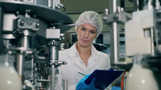 A woman checks bottles with milk, moving on a conveyor. Quality control of production line at a factory. Female worker controls the quality of production equipment.