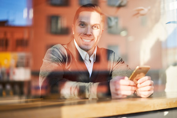 Happy man with phone in hand sitting in cafe, reflection of building in glass