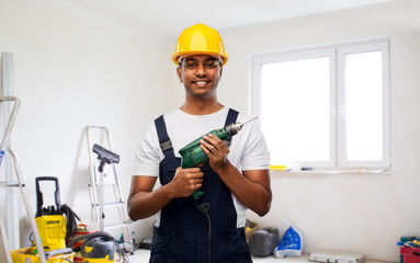 profession, building and repair concept - happy smiling indian repairman or builder in helmet with electric drill or perforator over empty room with construction equipment background