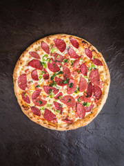 Pizza with ham, smoked sausage, bell pepper and cheese lies on a dark background. View from above