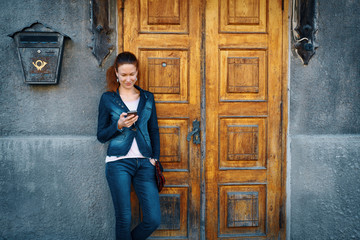 Young smiling woman denim clothing standing over old fashioned historical wooden door, rusty postbox and grey concrete wall background and texting on her phone on clear day. Travels in details concept