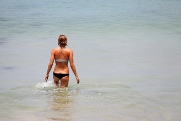 Fototapeta na wymiar Girl in bikini going to swim in the sea, splashing in water. Beach vacation on tropical sandy coast, slim female body, colorful seascape