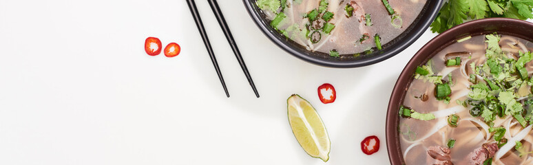 top view of pho in bowl near chopsticks, lime, chili and soy sauces and coriander on white...