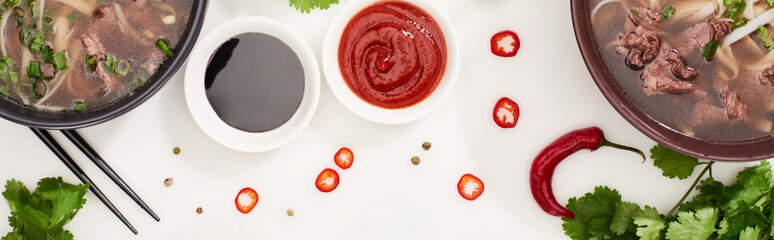 top view of pho in bowls near chopsticks, chili and soy sauces and coriander on white background, panoramic shot