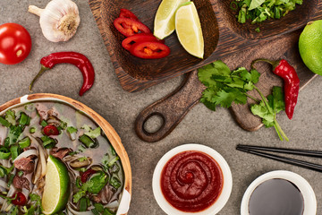 top view of pho in bowl near ingredients, chili and soy sauces and chopsticks on grey background