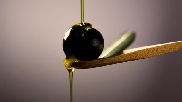 Olive oil is poured onto an olive lying on a spoon. Slow motion. Close-up of black olives on a wooden spoon on a gray background