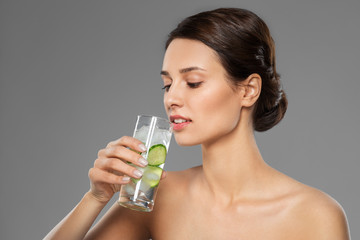 beauty and detox concept - woman drinking fresh water with cucumber and ice over grey background