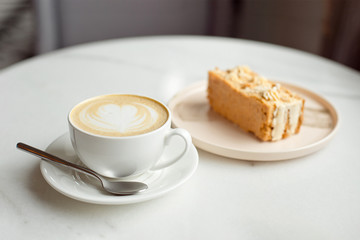 Slice of caramel cake and a fork on the right side. A cup of hot coffee with ground in the background.Close-up.