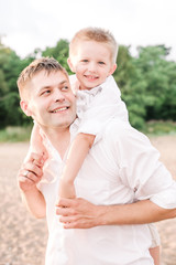 Fototapeta premium Happy dad carries on the back of his smiling son. Father and son walk on the beach, dressed in bright clothes. Vertical format.