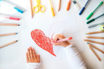 Close up of child's hands drawing red heart at white paper within colorful pens and pencils.
