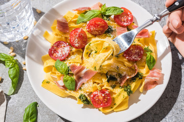 partial view of woman eating tasty Pappardelle with tomatoes, pesto and prosciutto near water on grey surface