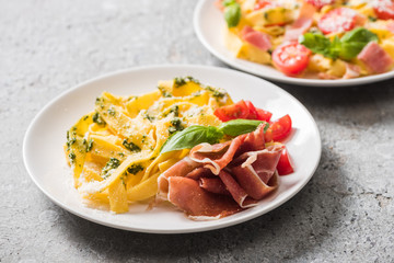 selective focus of cooked Pappardelle with tomatoes, basil and prosciutto near ingredients on grey surface