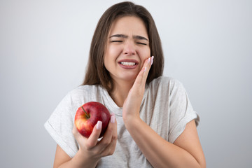 young brunette woman suffering toothache holding red apple in her hand standing on isolated white...