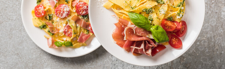 selective focus of cooked Pappardelle with tomatoes, basil and prosciutto on plates on grey surface, panoramic shot