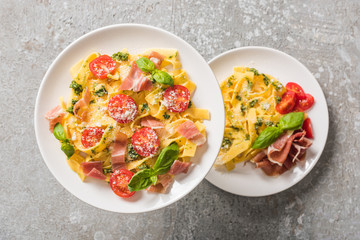 selective focus of cooked Pappardelle with tomatoes, basil and prosciutto on plates on grey surface