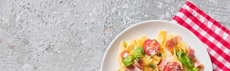 top view of cooked Pappardelle with tomatoes, basil and prosciutto on plaid napkin on grey surface, panoramic shot