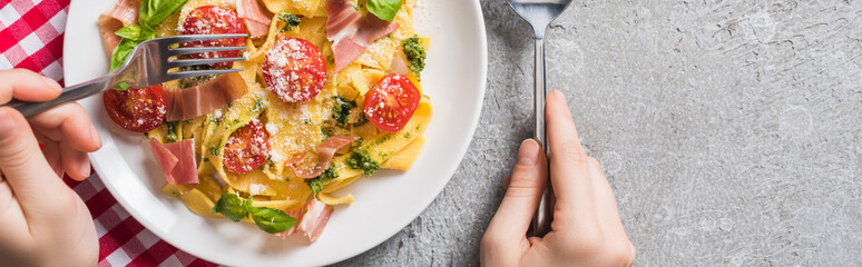 partial view of woman eating Pappardelle with tomatoes, basil and prosciutto on plaid napkin on grey surface, panoramic shot