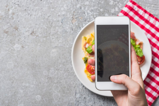 partial view of woman taking picture of Pappardelle with tomatoes, basil and prosciutto on smartphone on grey surface