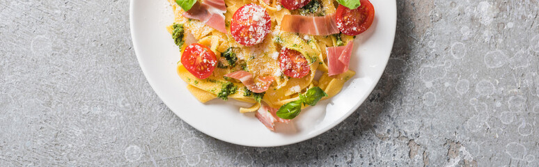 top view of cooked Pappardelle with tomatoes, pesto sauce and prosciutto on grey surface, panoramic shot