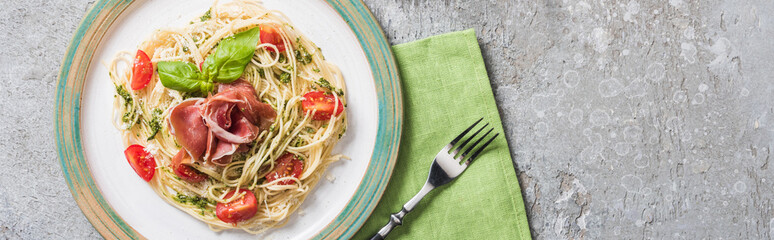 top view of cooked Pappardelle with tomatoes, basil and prosciutto on plate on green napkin with fork on grey surface, panoramic shot