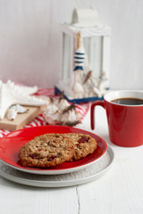 Oatmeal cookies with cranberries lie on a red plate, next to a cup of black coffee, a flashlight, seashells and an old notebook. The composition is on a striped tablecloth and white wooden background.