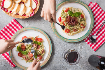 cropped view of women eating Pappardelle with tomatoes, basil and prosciutto near baguette, red wine on grey surface